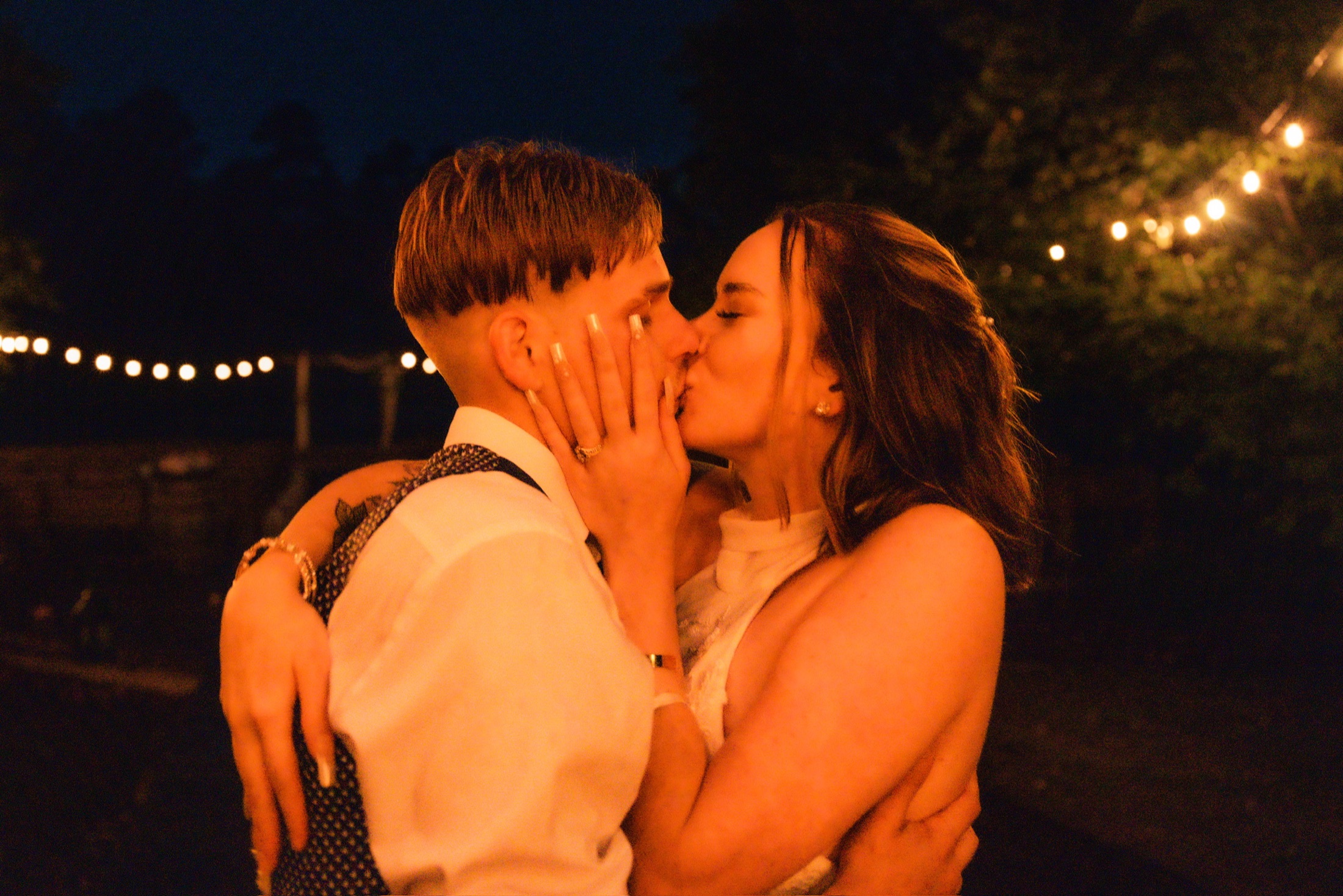 Newlyweds kissing beneath warm string lights at night